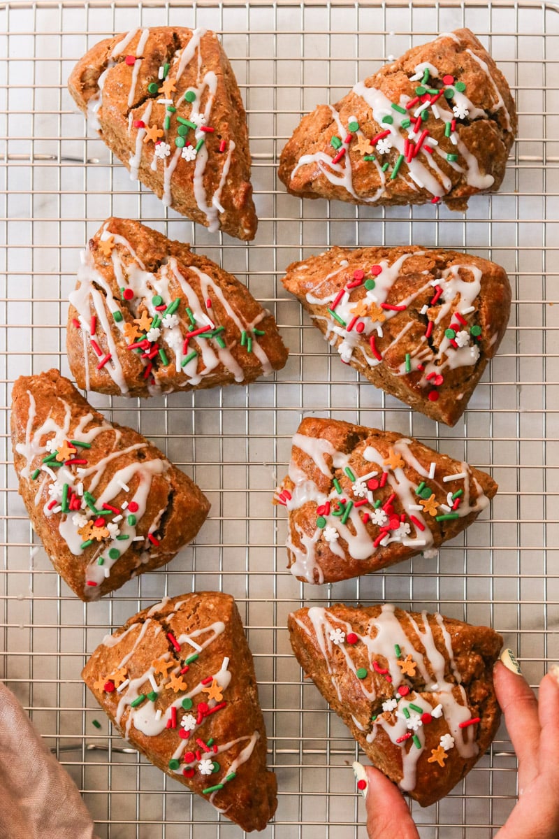 Glazed gingerbread scones topped with festive sprinkles on a wire rack, with a hand lifting one.