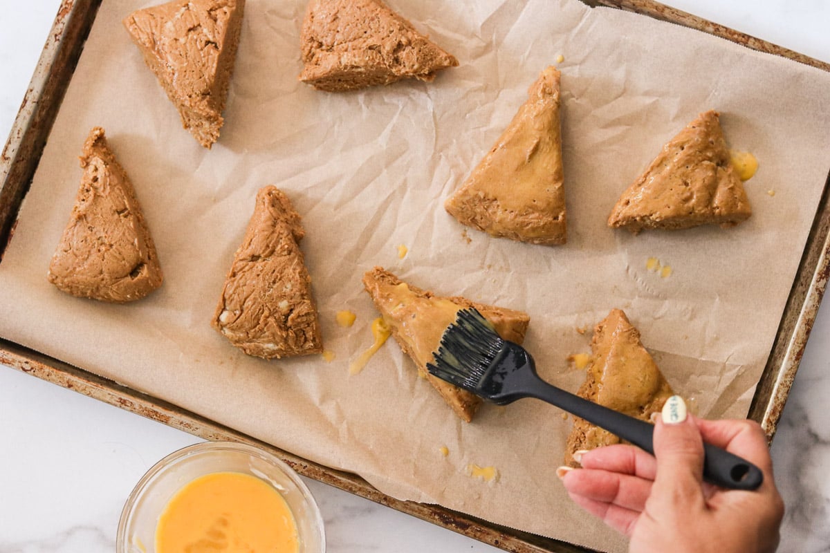 Chilled scone dough wedges on a parchment-lined pan being brushed with egg wash.