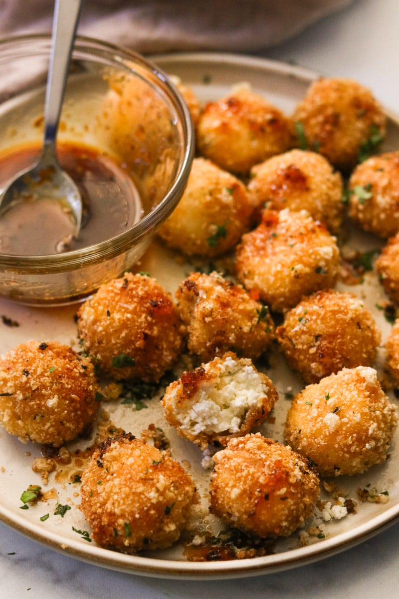 Side view of fried goat cheese balls on a plate, drizzled with honey and topped with flaky sea salt and chopped parsley; a bite is taken out of one.