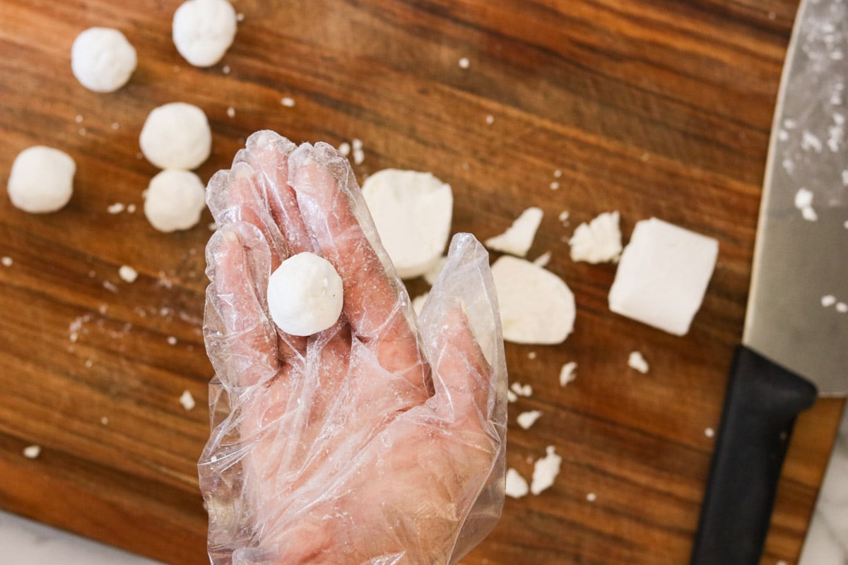Gloved hand holding a rolled goat cheese ball over a wooden board with more goat cheese balls.
