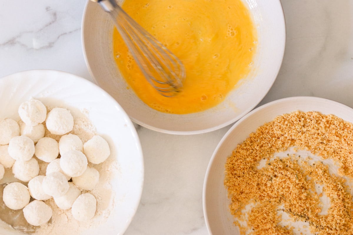 Three shallow bowls holding beaten egg, seasoned panko crumbs, and rolled goat cheese balls.