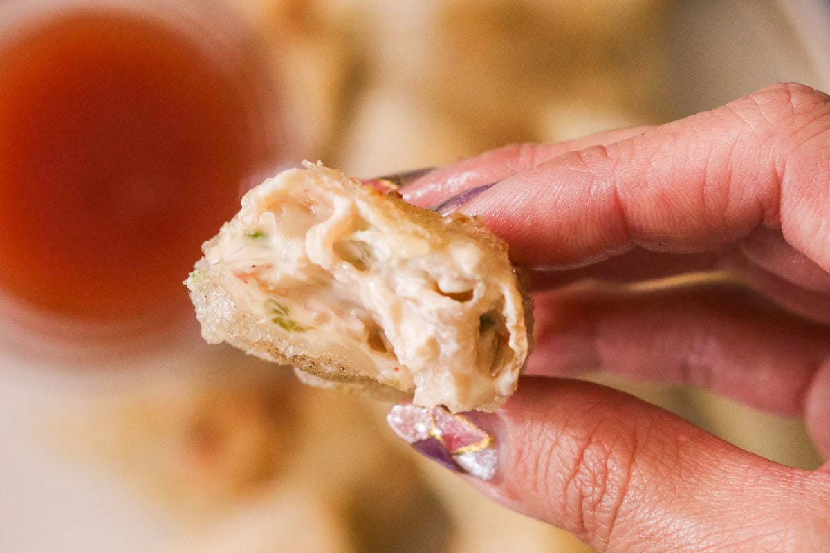 Hand holding a gluten-free crab rangoon with a bite taken out, showing the creamy filling inside.