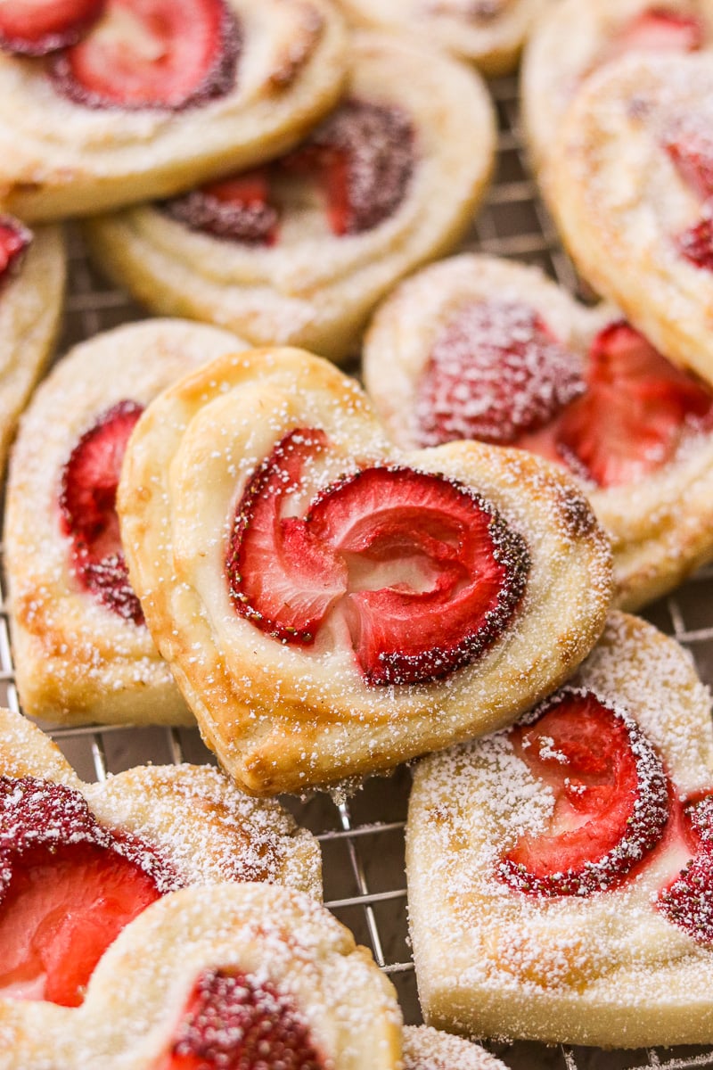 Close-up of heart-shaped gluten-free danishes on a wire cooling rack, each topped with a strawberry center and dusted with powdered sugar.
