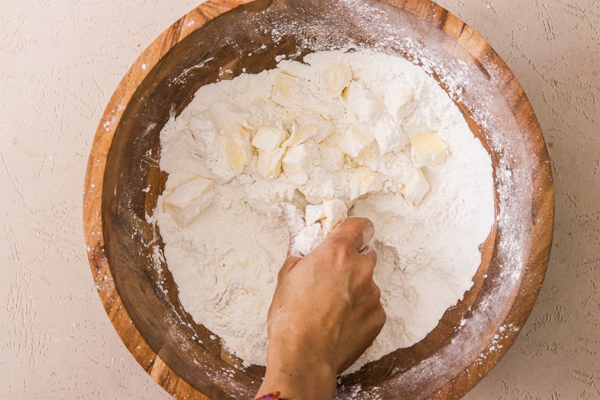Gluten-free pie crust mix and cold butter being mixed by hand in a large wooden bowl until crumbly.