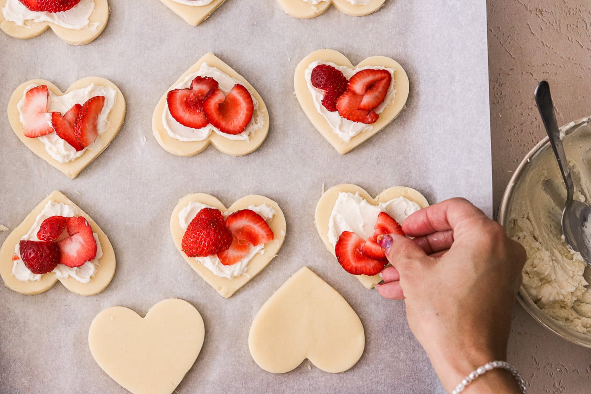 Heart-shaped dough topped with cream cheese filling and fresh strawberry slices before baking.