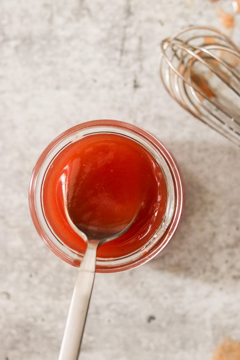 Jar of homemade sweet and sour sauce with a spoon inside and a whisk resting beside it