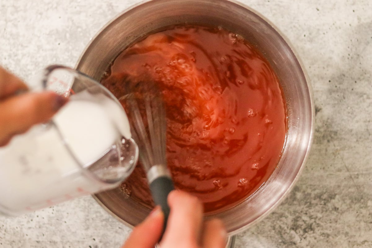Cornstarch slurry being poured into boiling sweet and sour sauce while whisking until thick and glossy