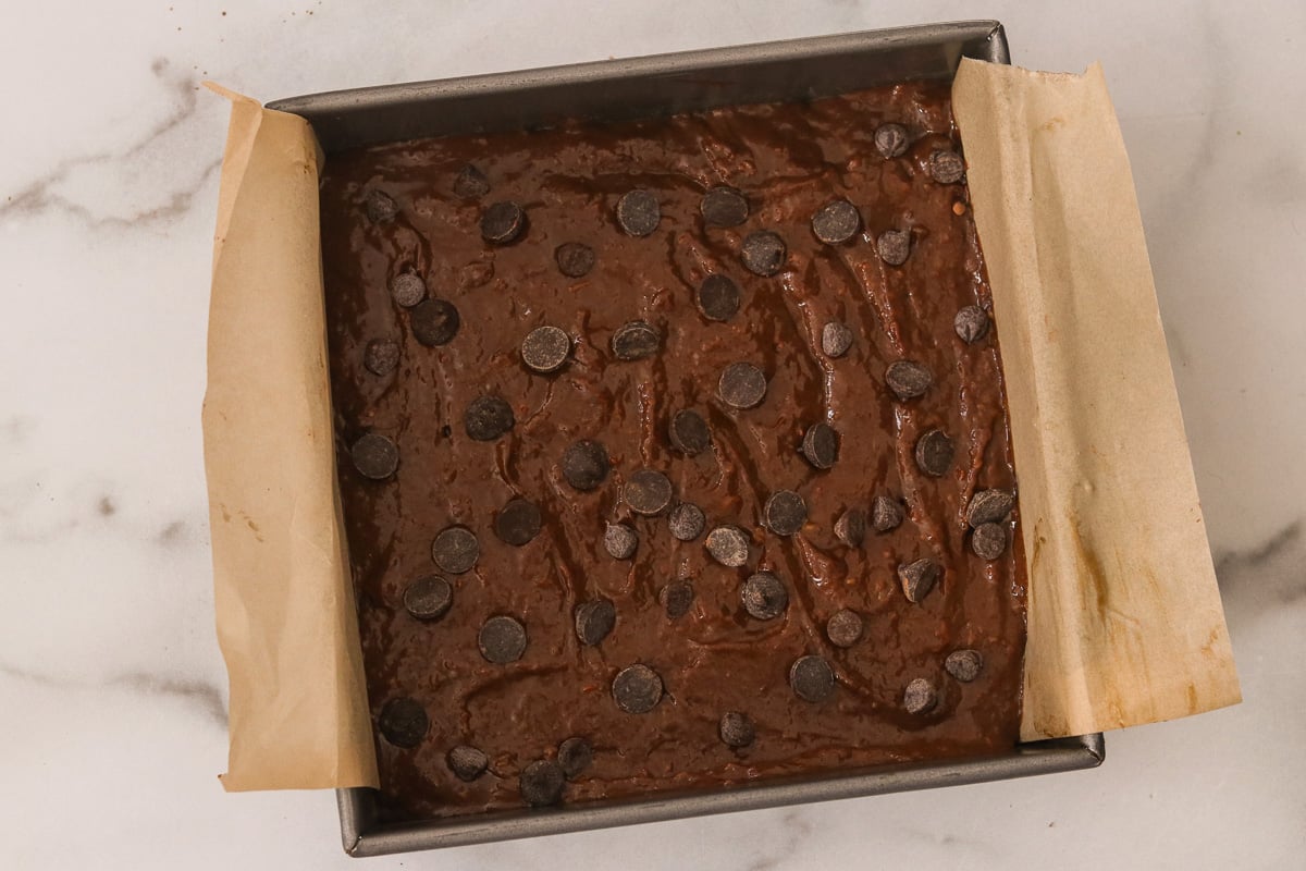 Sweet potato brownie batter in a parchment-lined pan spread out evenly. Remaining chocolate chips are sprinkled over the top.
