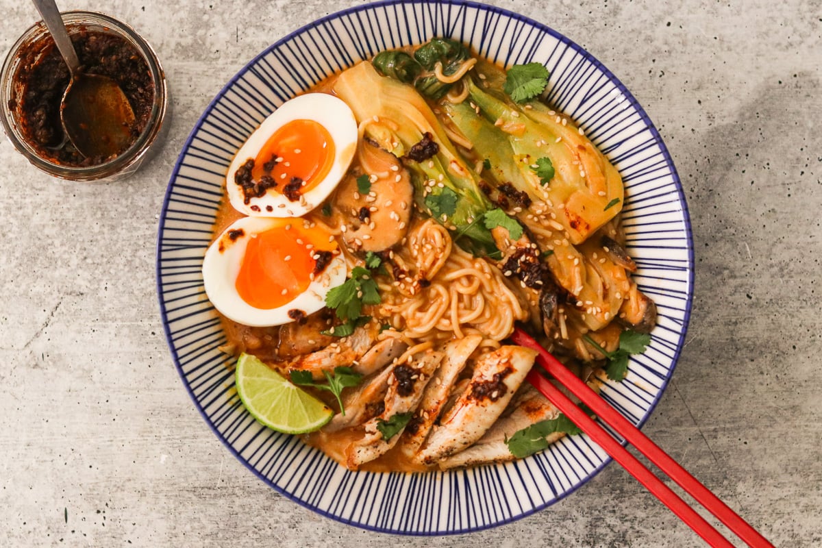 Coconut curry ramen in a blue and white striped bowl topped with cilantro, lime wedges, and soft-boiled eggs, with red chopsticks resting inside.