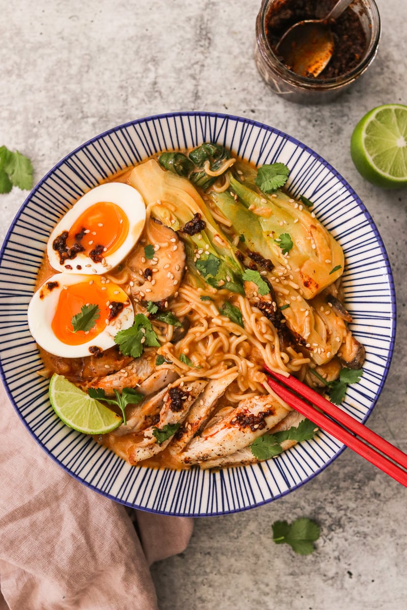 Coconut curry ramen in a blue and white striped bowl topped with cilantro, lime wedges, and soft-boiled eggs, with red chopsticks resting inside.
