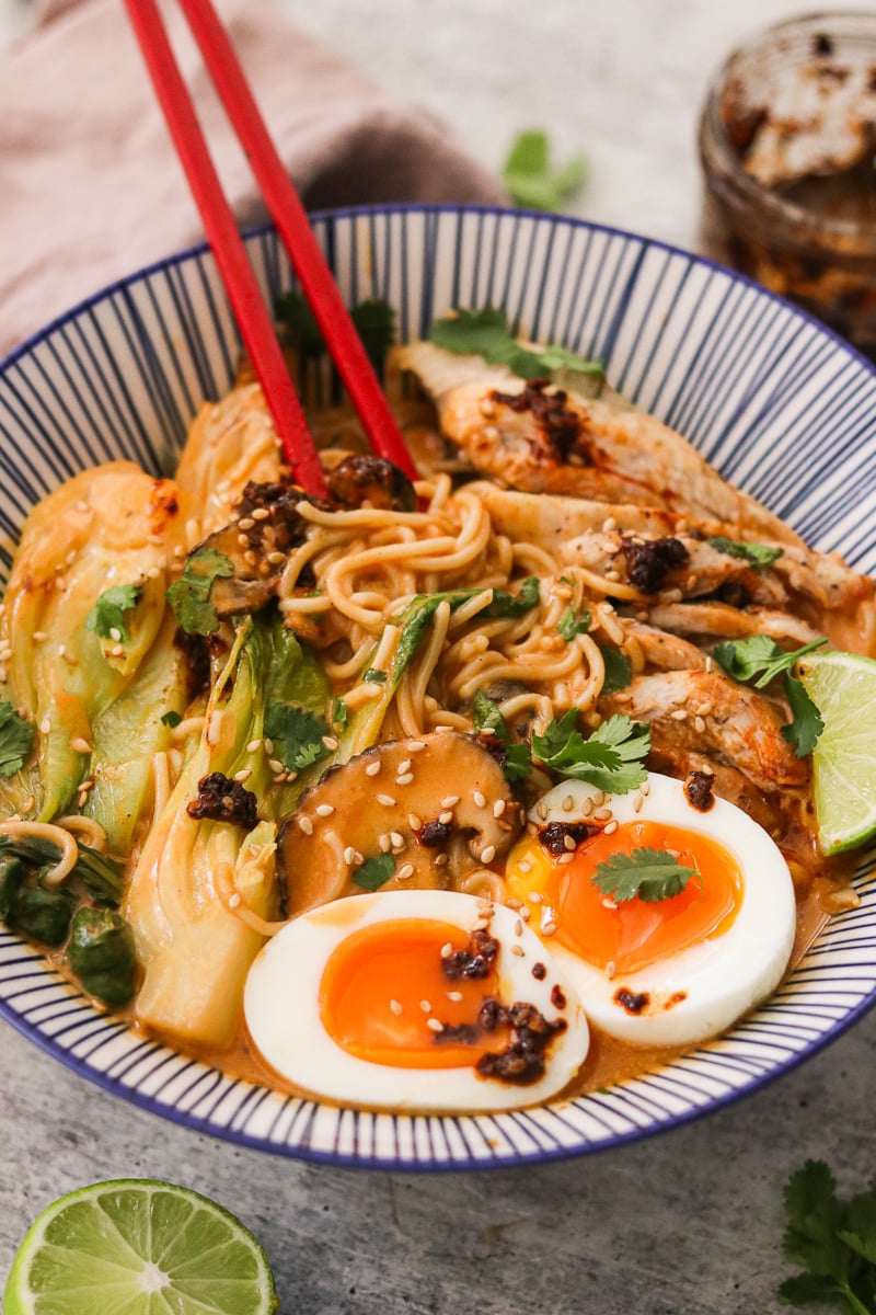 Close-up of coconut curry ramen in a blue and white striped bowl with cilantro, lime wedges, and soft-boiled eggs as red chopsticks lift a bite.