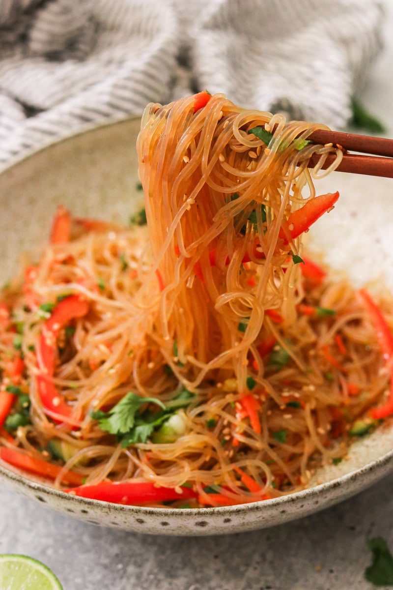 Sesame noodle salad garnished with cilantro and sesame seeds being lifted from a bowl with chopsticks.