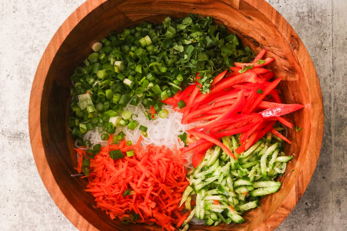 Cooled vermicelli noodles in a large wooden bowl topped with sliced red bell pepper, shredded carrot, cucumbers, green onions, and fresh cilantro.