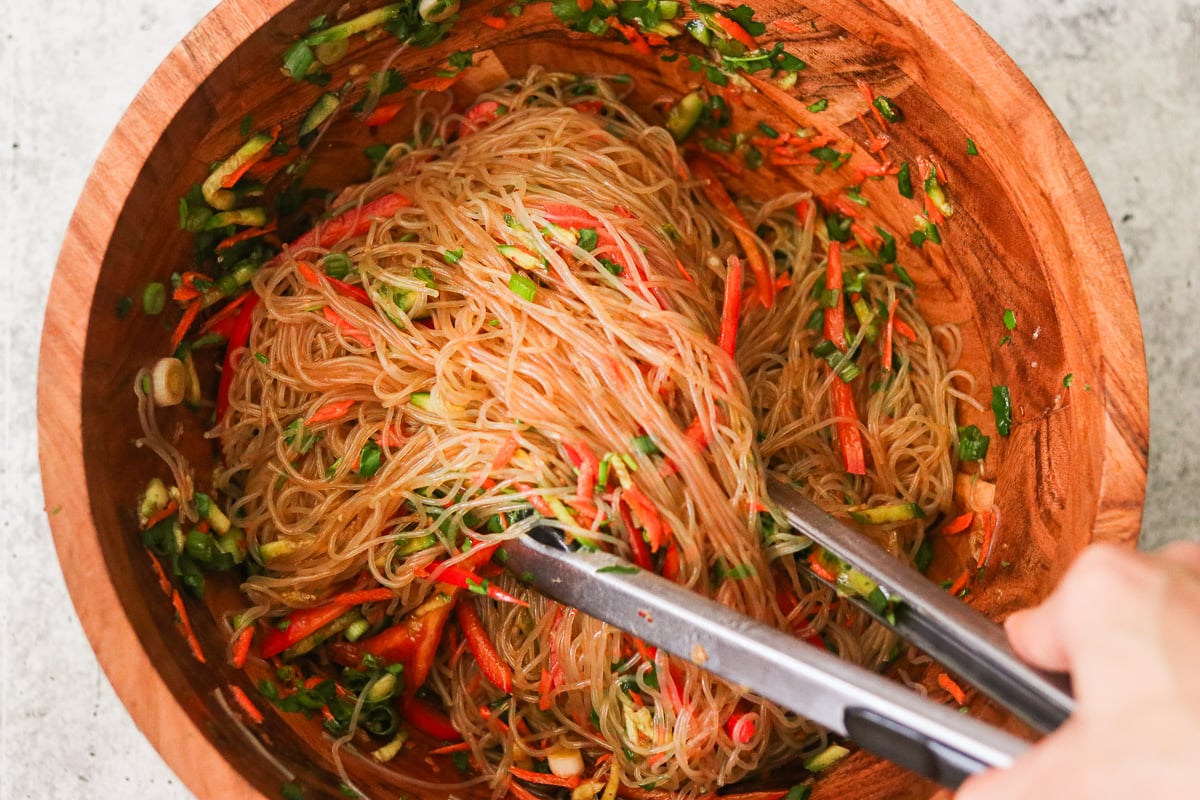 Sesame dressing poured over vermicelli noodles and fresh vegetables in a large bowl while tongs lift and toss the salad to coat evenly.