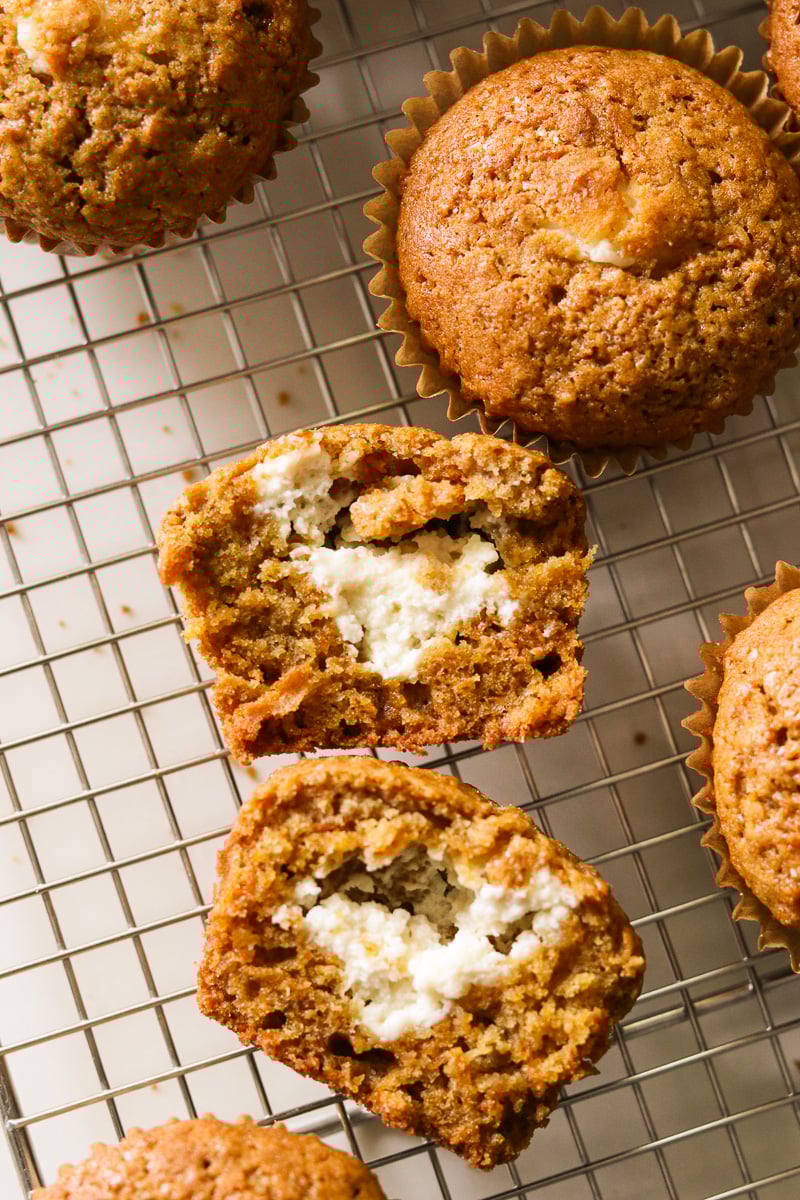 Close-up of a gluten-free carrot cake muffin on its side, showing the creamy cream cheese filling inside, resting on a wire rack with other muffins in the background.