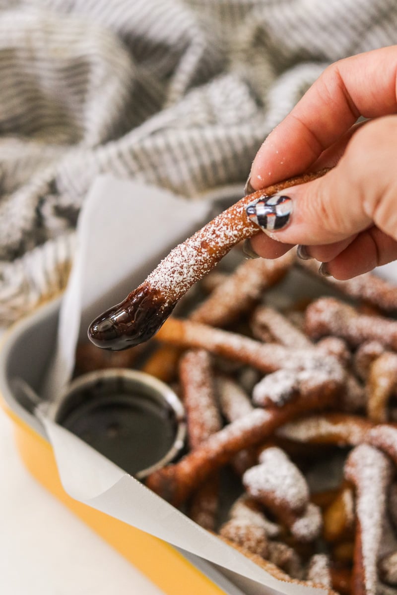Funnel cake fry dipped in chocolate sauce being held, with a tray of funnel cake fries and chocolate dipping sauce in the background.