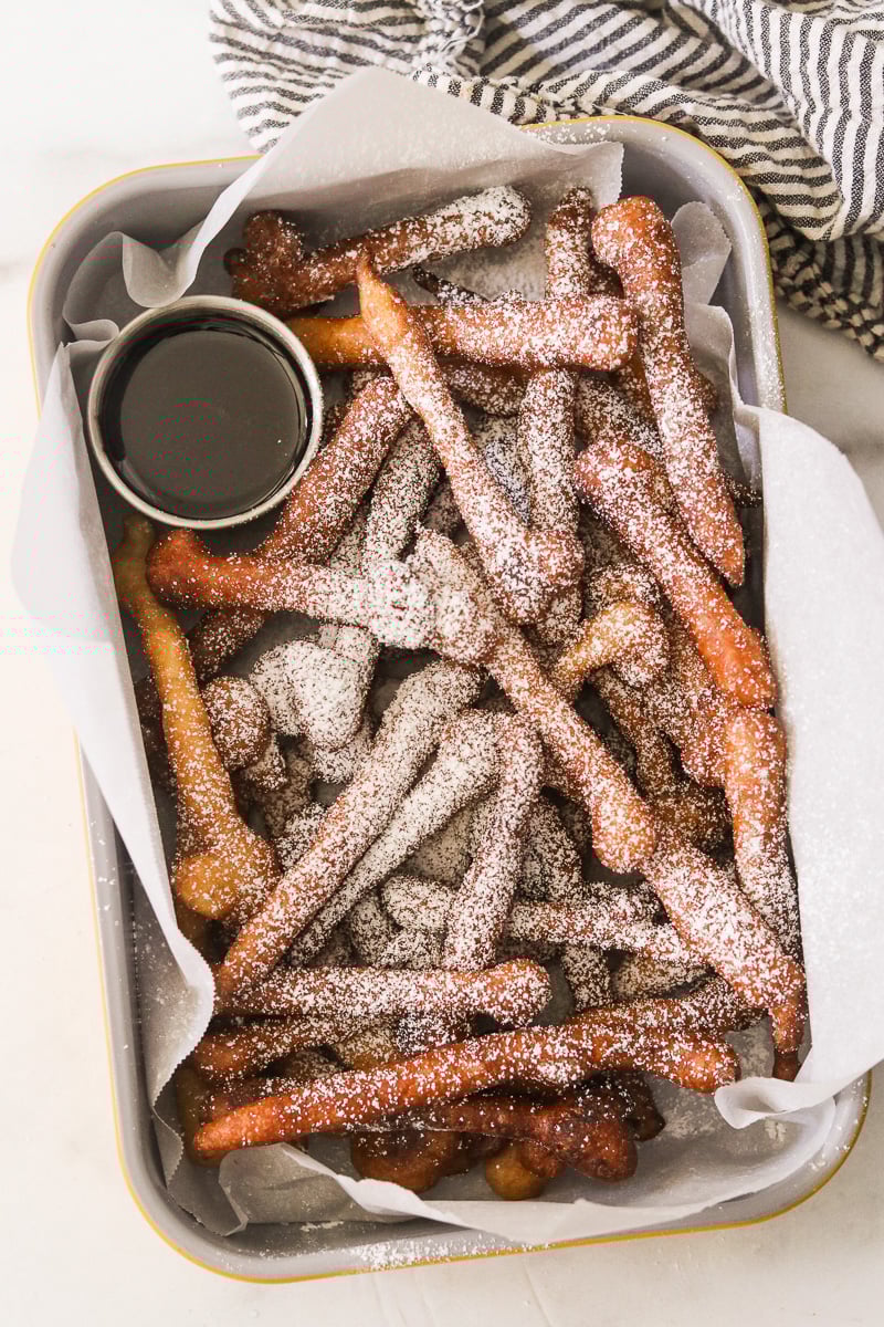 Funnel cake fries dusted with powdered sugar in a baking dish served with a bowl of chocolate dipping sauce.