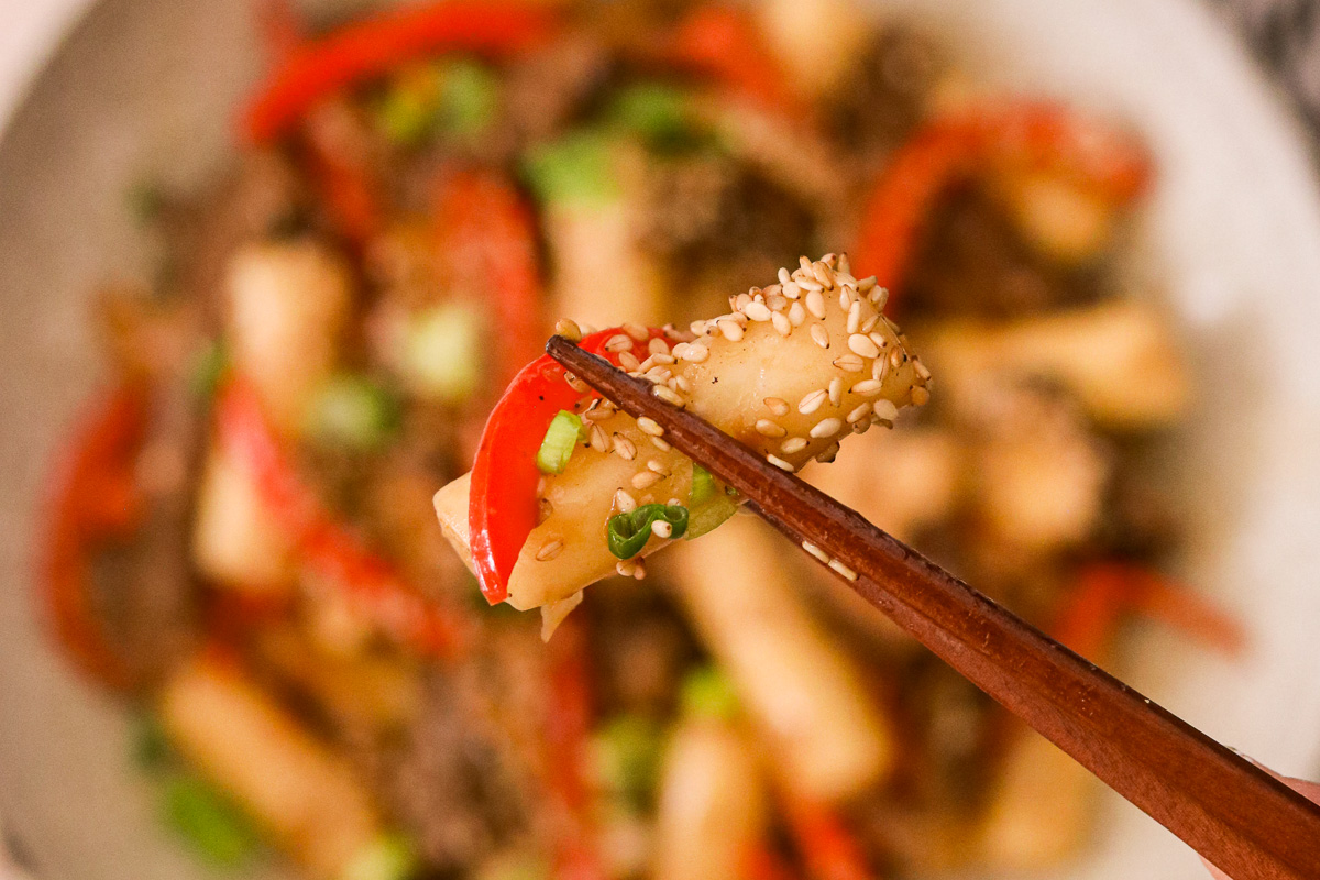 chopsticks holding up a korean rice cake with in sauce and topped with sesame seeds.