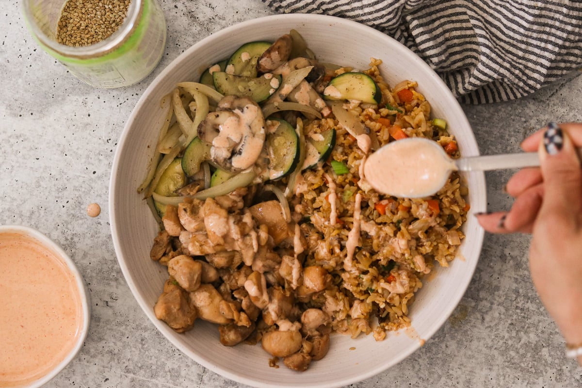 Hibachi chicken in a bowl with fried rice and vegetables, being topped with yum yum sauce.