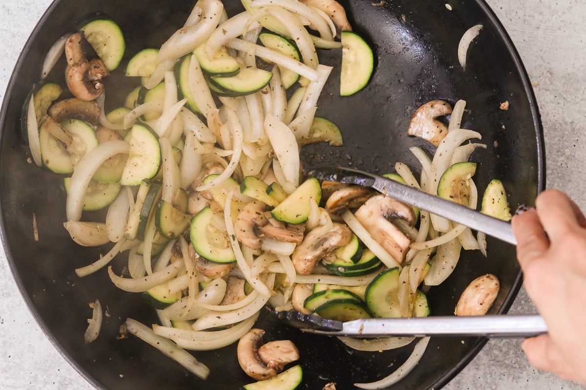Sliced onion, zucchini, and mushrooms cooking in avocado oil in a large skillet; tongs are being used to toss them.