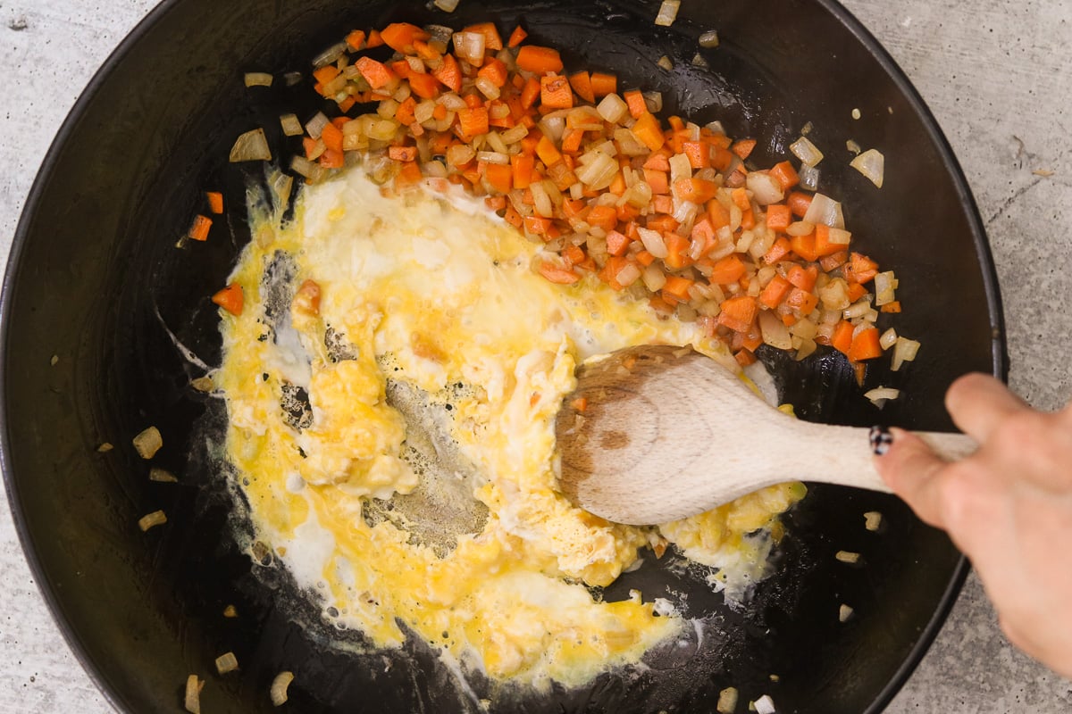 Eggs cracked into the skillet beside the vegetables being scrambled with a wooden spoon.