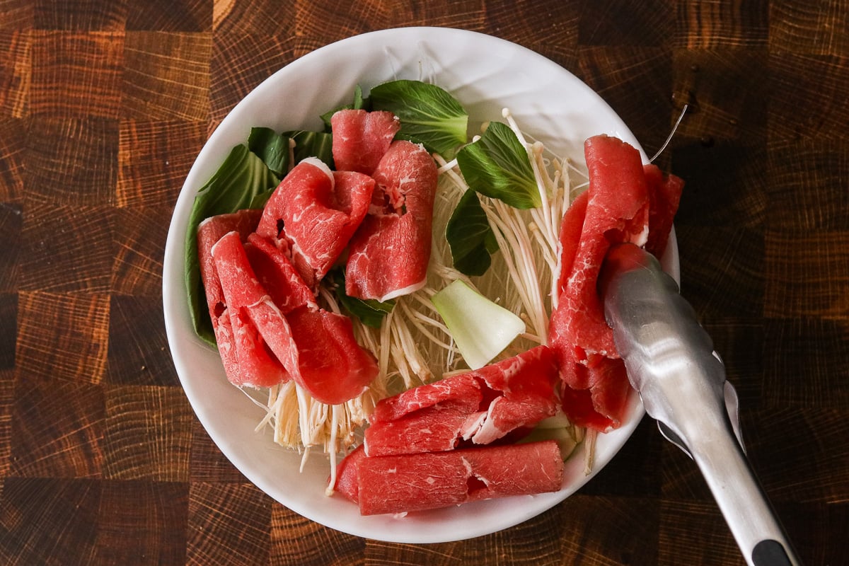 Vermicelli noodles, enoki mushrooms, bok choy, and thinly sliced beef layered in a large microwave-safe bowl.