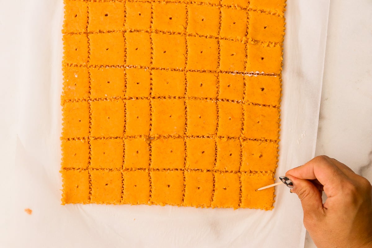 Rolled dough cut into small square crackers with crimped edges, each one being poked in the center with a toothpick to create a small hole before baking.