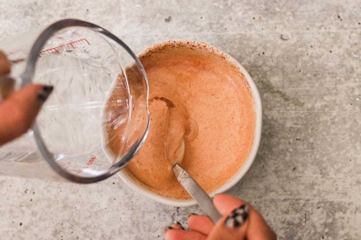 A glass measuring cup slowly pouring a small amount of water into the yum yum sauce.