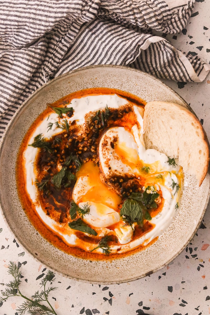 Turkish eggs recipe in a speckled bowl with crusty bread. A striped linen towel is next to the bowl.