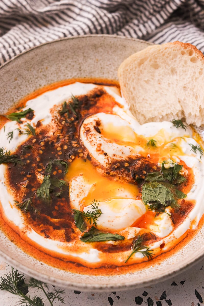 Close up of Turkish eggs with bread in a speckled bowl on a linen towel.
