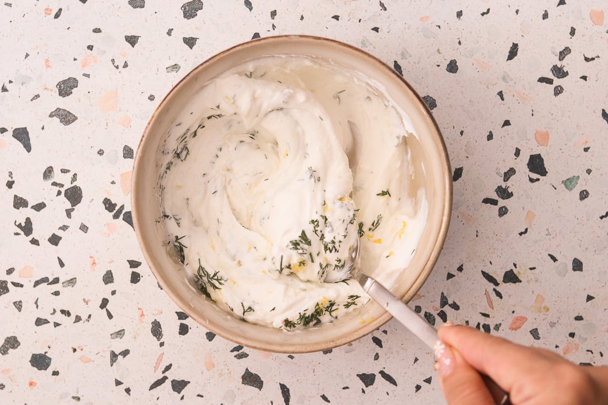 Greek yogurt being mixed with garlic, lemon zest, dill, and salt in a small bowl.