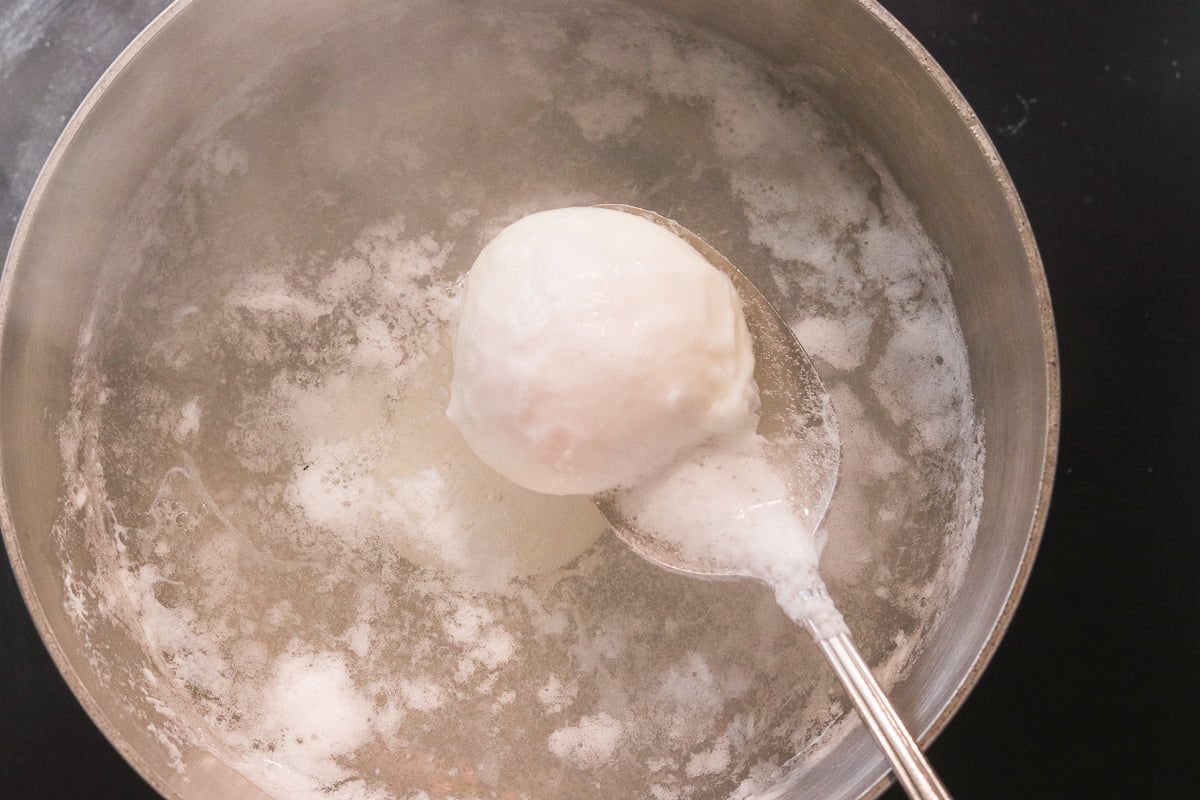 Poached egg being lifted with a spoon from a pot of gently simmering water.