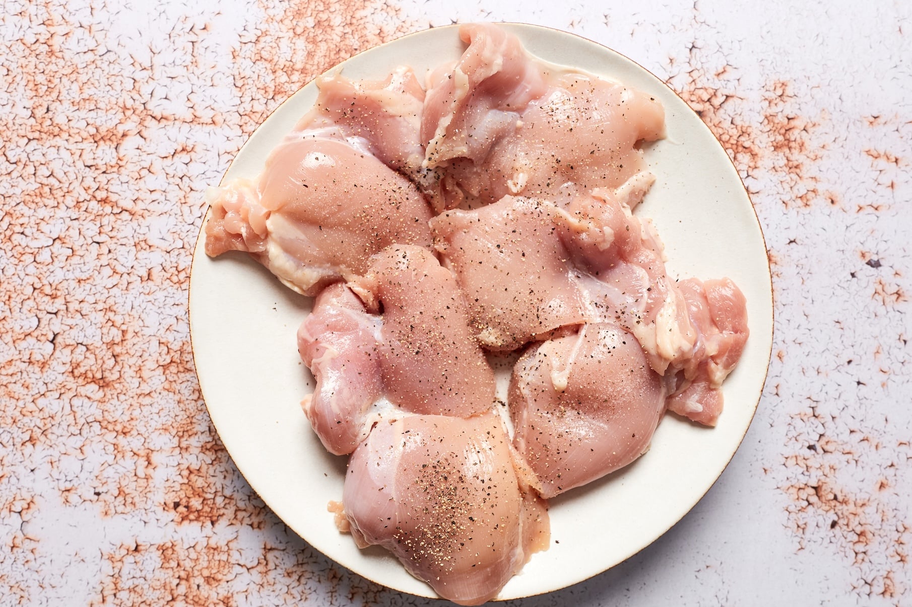 Raw chicken thighs on a plate seasoned generously with salt and black pepper before cooking.