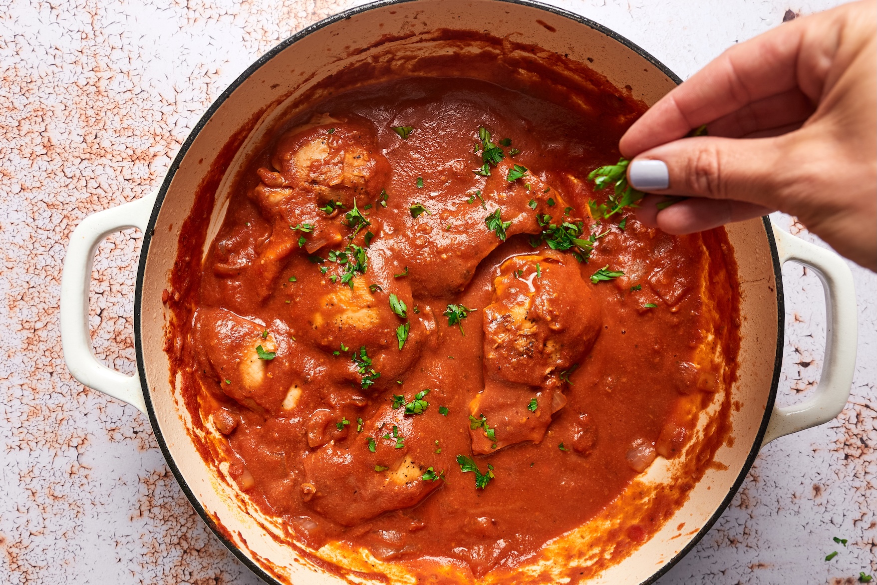 Creamy tomato chicken simmering in the skillet until the sauce thickens slightly and the chicken is fully cooked; a hand is garnishing the chicken with fresh basil.