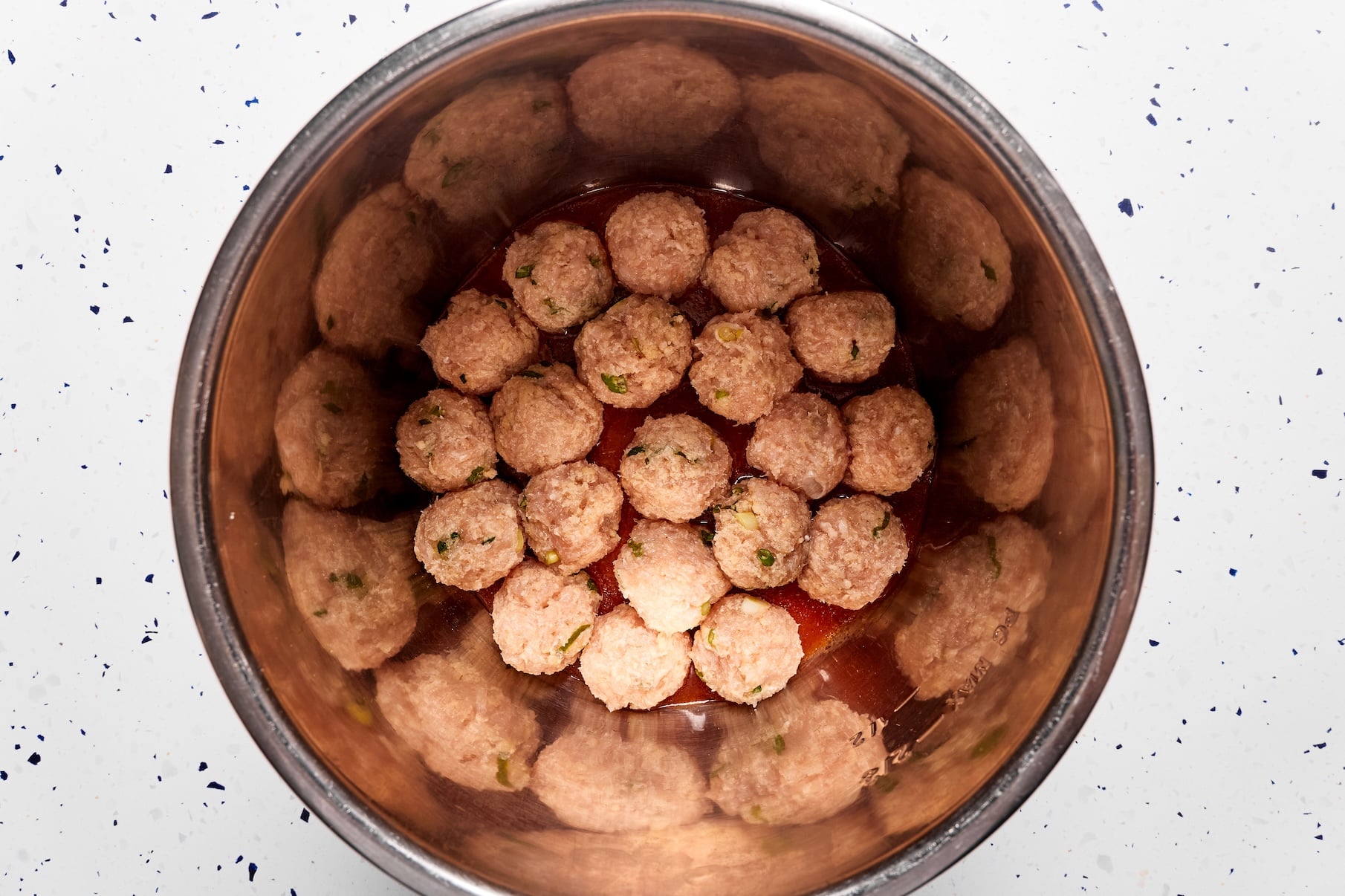 Buffalo sauce spread in the bottom of the Instant Pot with raw chicken meatballs arranged evenly on top before cooking.