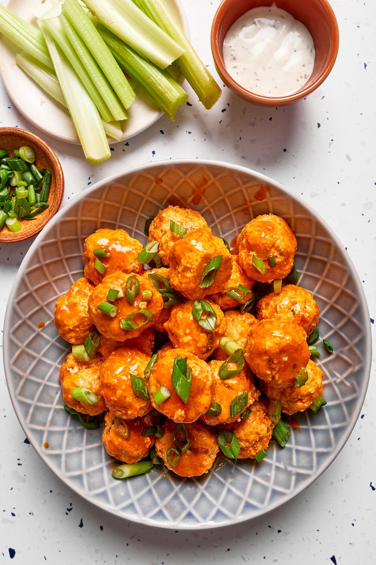Buffalo chicken meatballs in a small bowl coated in buffalo sauce and garnished with sliced green onions, with celery sticks, ranch dressing, and extra chopped green onions sitting behind the bowl.