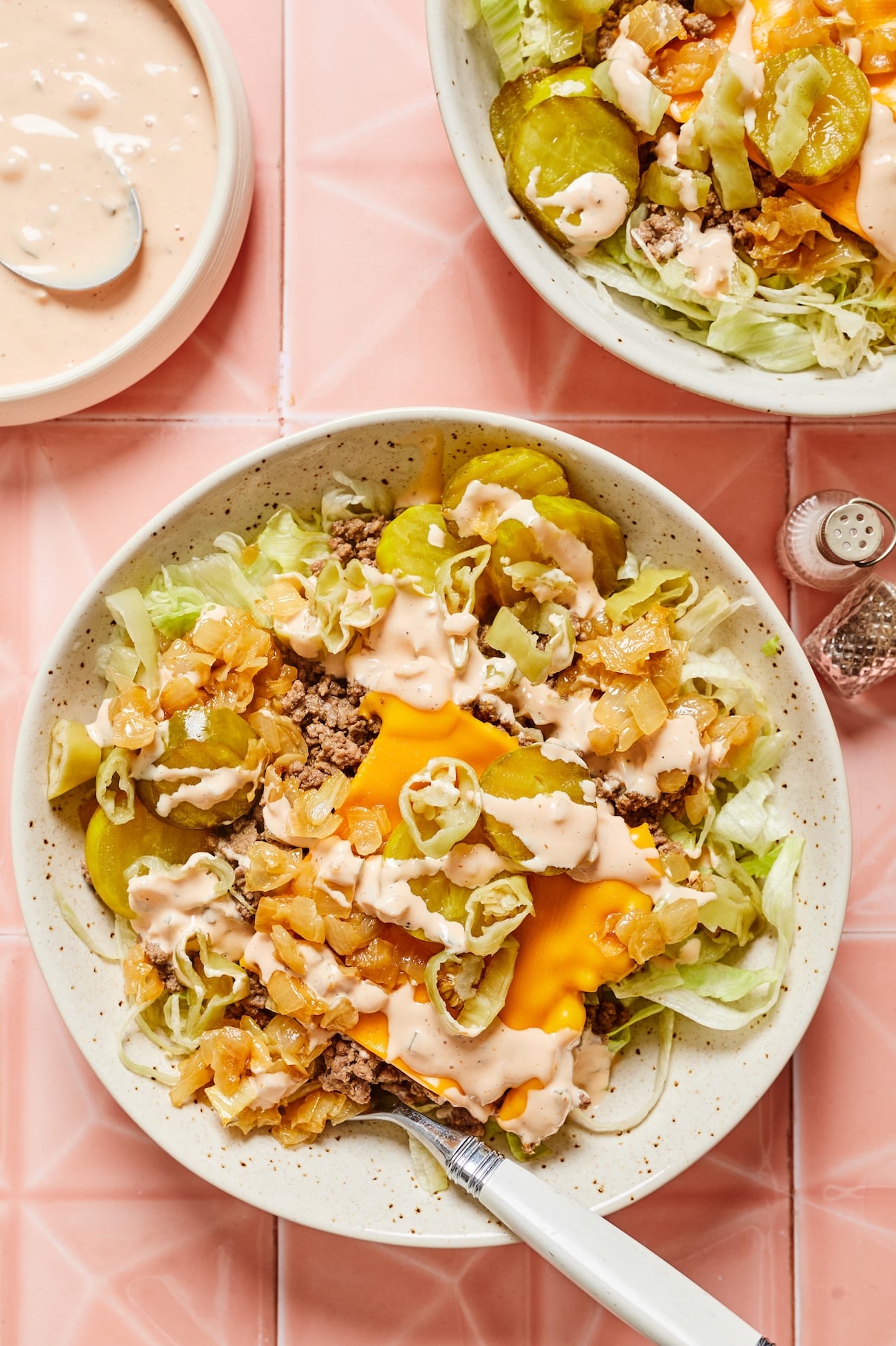 Two bowls of cheeseburger salad topped with ground beef, cheddar cheese, caramelized onions, pickles, pepperoncini peppers, and animal sauce, with extra sauce in a bowl to the side.