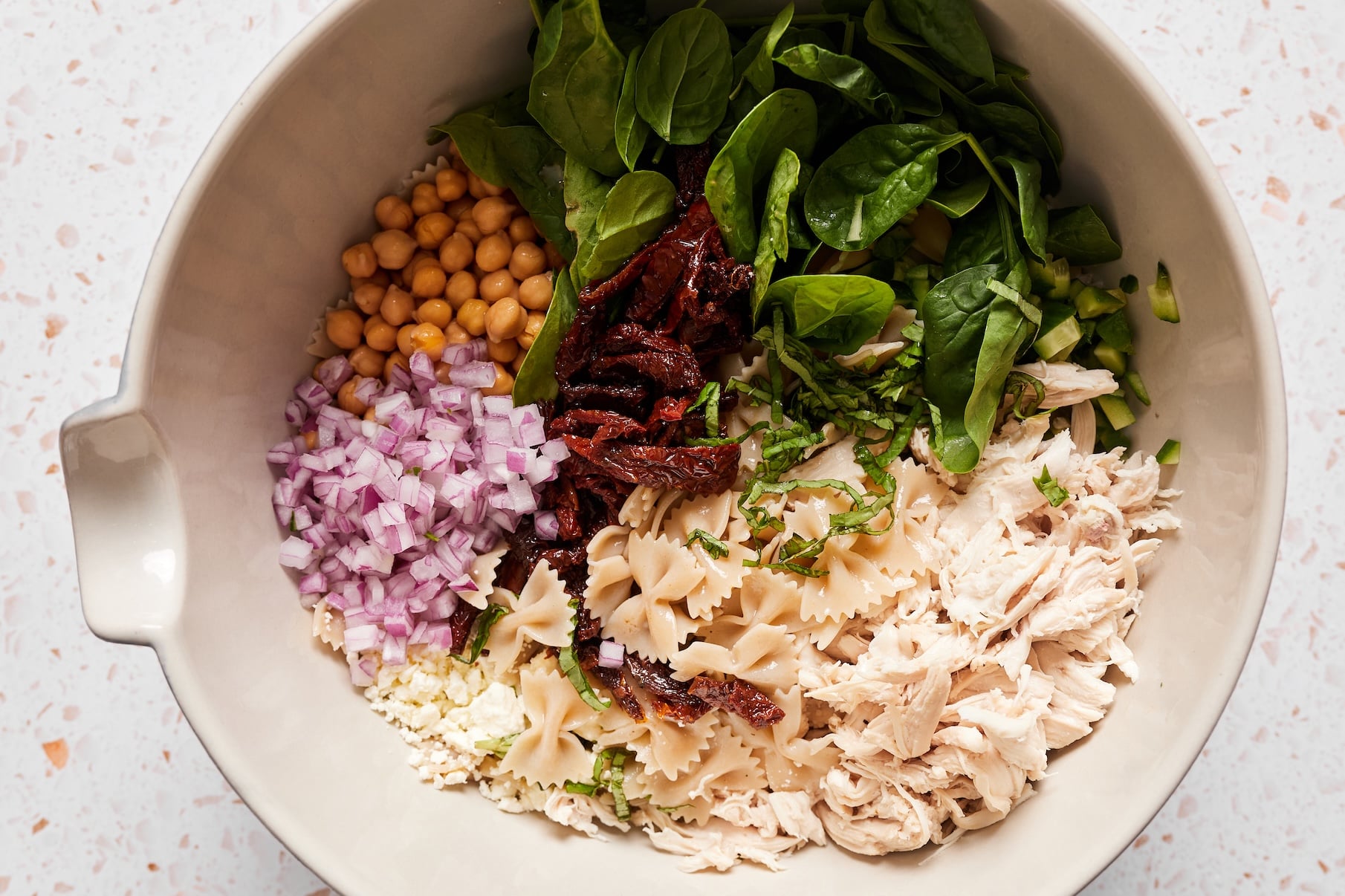 Cooked pasta, chickpeas, shredded chicken, sun-dried tomatoes, baby spinach, cucumber, red onion, feta, and basil in a large serving bowl.