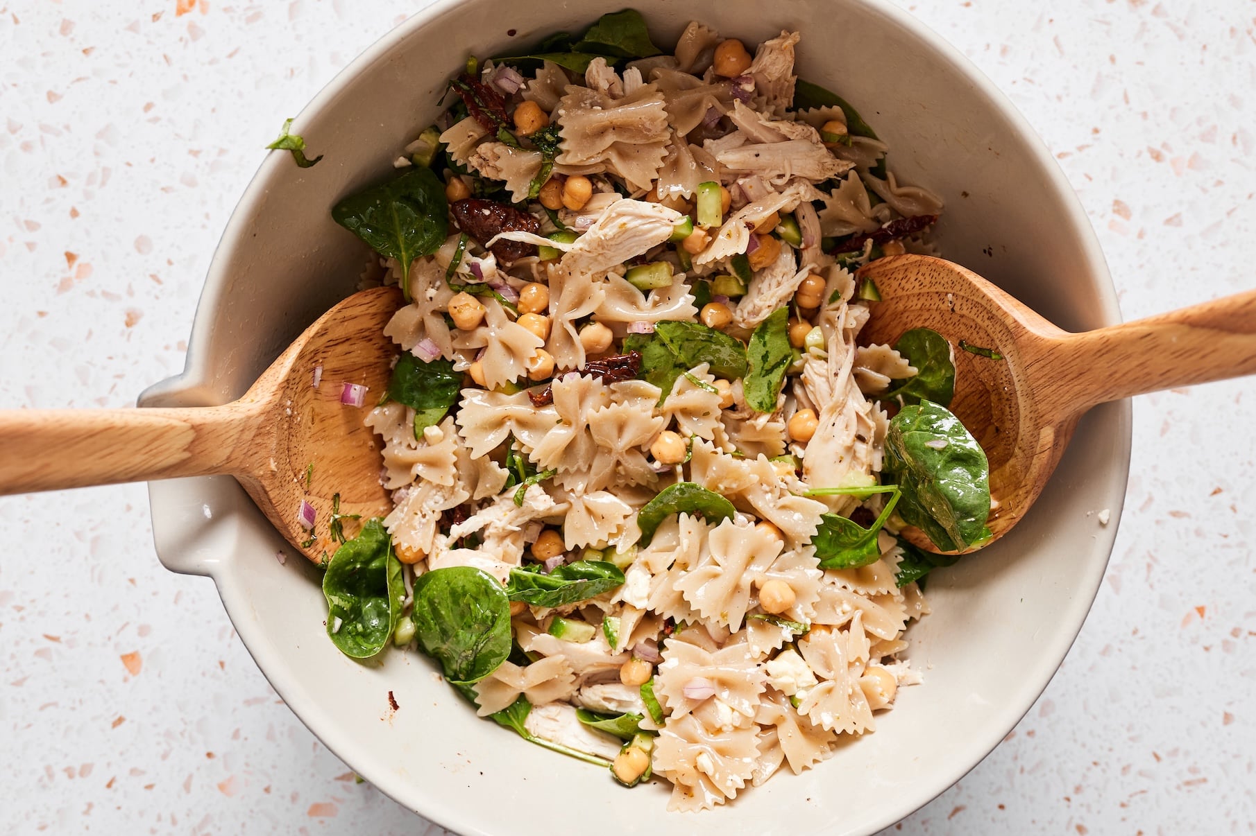 Two wooden salad spoons tossing chickpea pasta salad with chicken, vegetables, feta, and homemade dressing until everything is evenly coated.