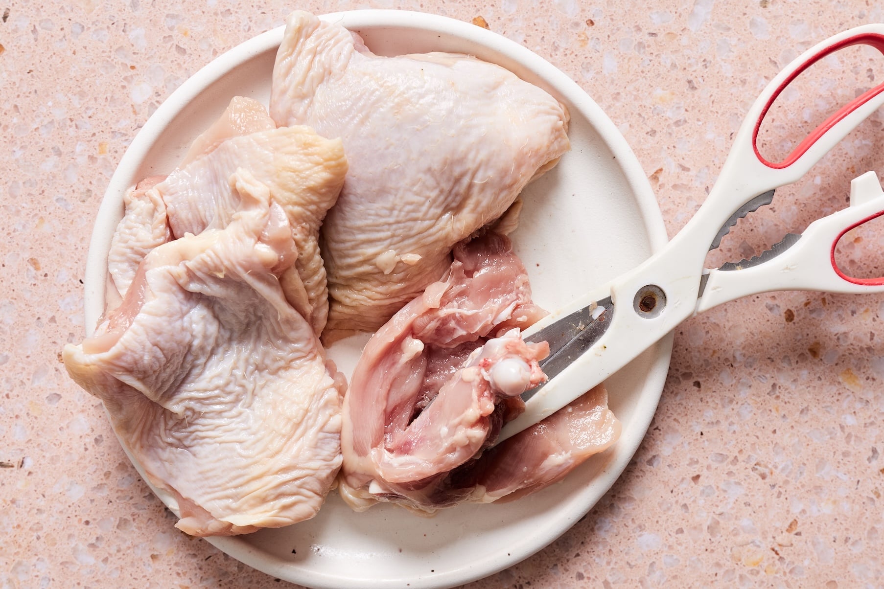 Raw chicken thighs on a white plate with scissors cutting into one.