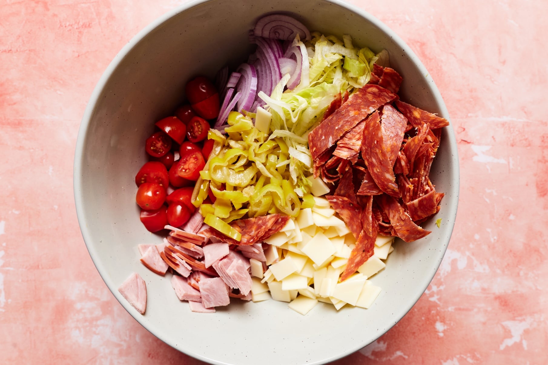 Shredded lettuce, tomatoes, red onion, pepperoncini, ham, salami, and provolone in a large mixing bowl.