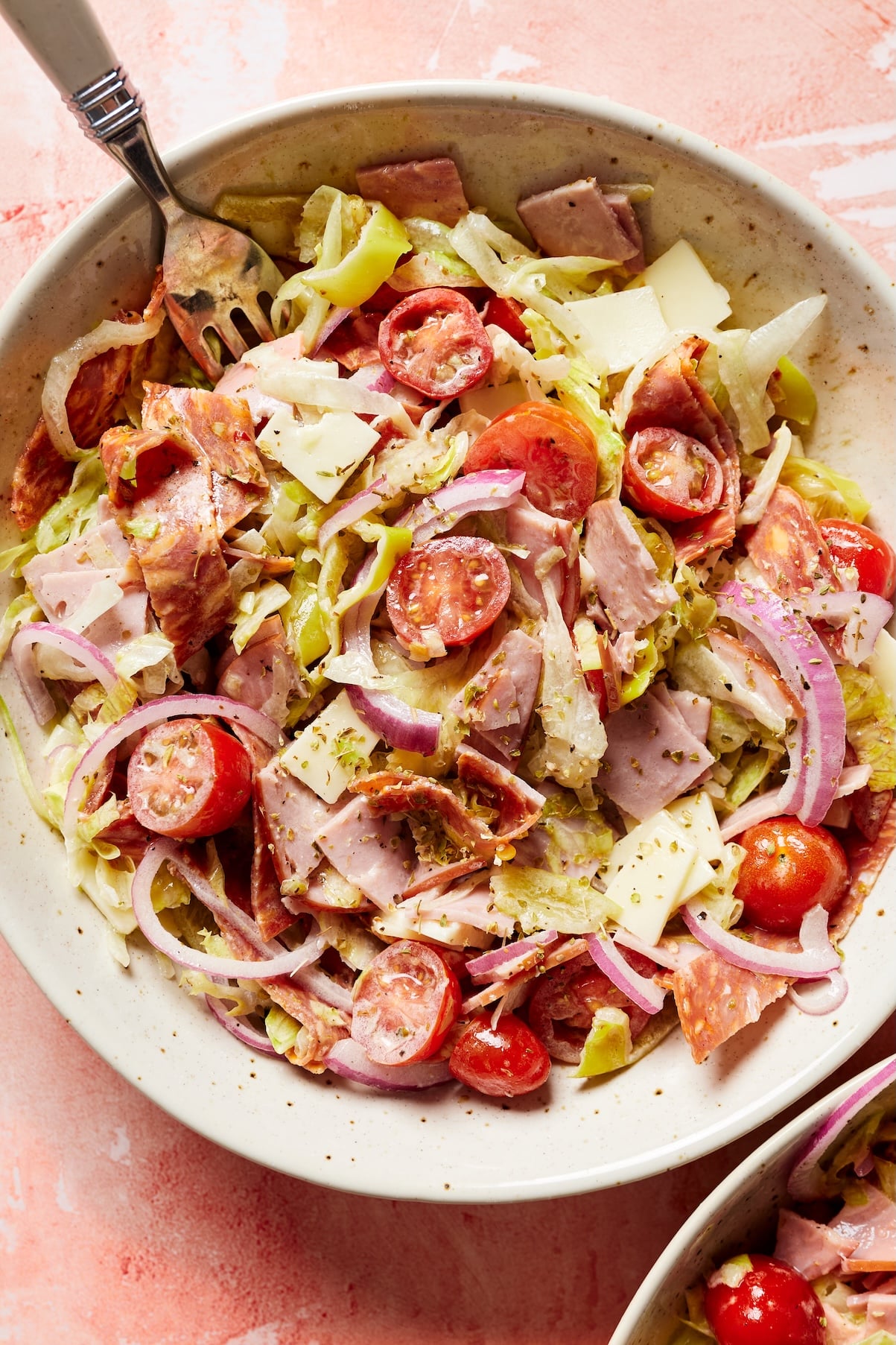 Close-up of a bowl of sub in a tub with crisp lettuce, deli meats, provolone, tomatoes, and pepperoncini, with a fork resting inside.