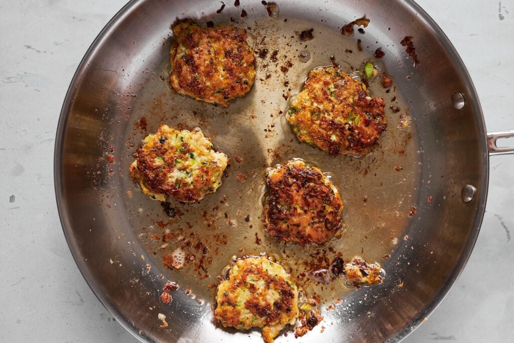 Five golden-brown, crispy chicken zucchini fritters arranged in a large skillet.