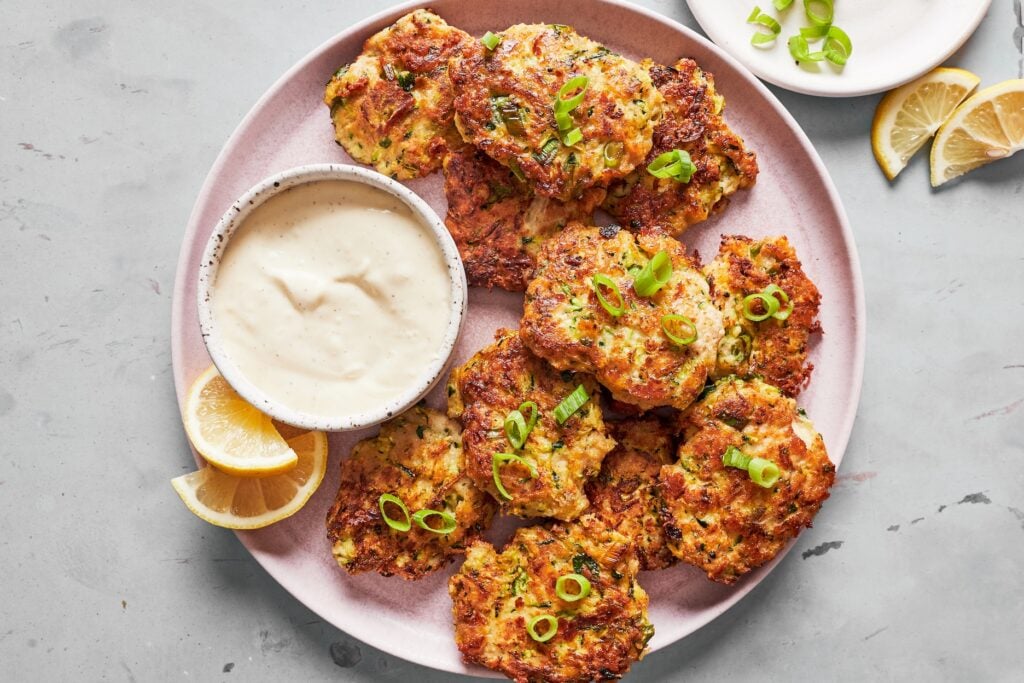 Gluten-free zucchini fritters on a pink plate with green onions, creamy garlic aioli for dipping, and two lemon wedges.