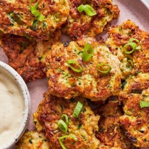 Zucchini fritters topped with green onions served on a pink plate with creamy garlic aioli.