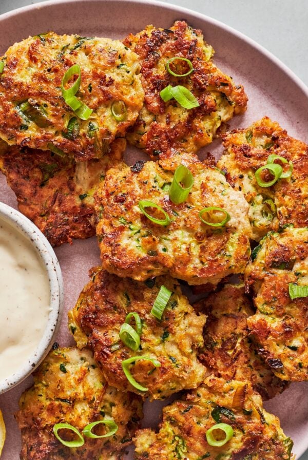 Zucchini fritters topped with green onions served on a pink plate with creamy garlic aioli.