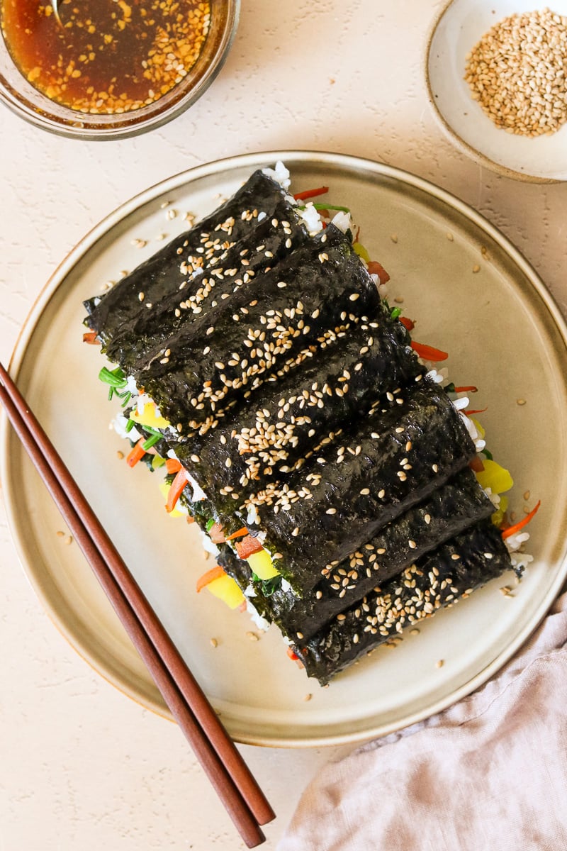 Mini kimbap rolls garnished with sesame seeds and stacked on a plate, with a small glass bowl of dipping sauce in the background.
