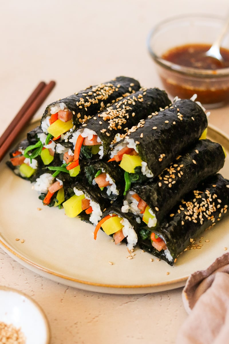 Mini kimbap rolls garnished with sesame seeds and stacked on a plate, with a small glass bowl of dipping sauce in the background.