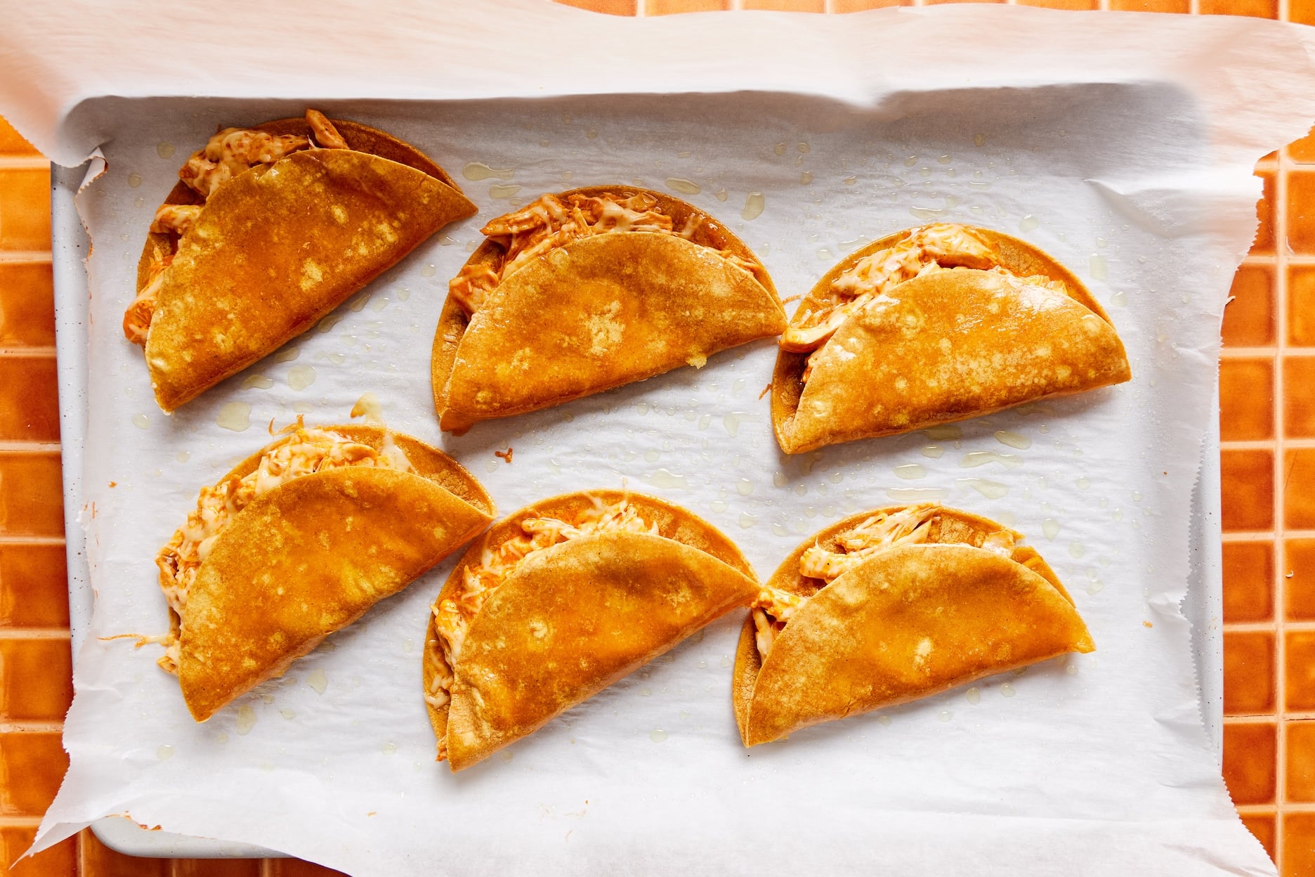 Six crispy and golden buffalo chicken tacos on a parchment-lined baking sheet after the first bake.