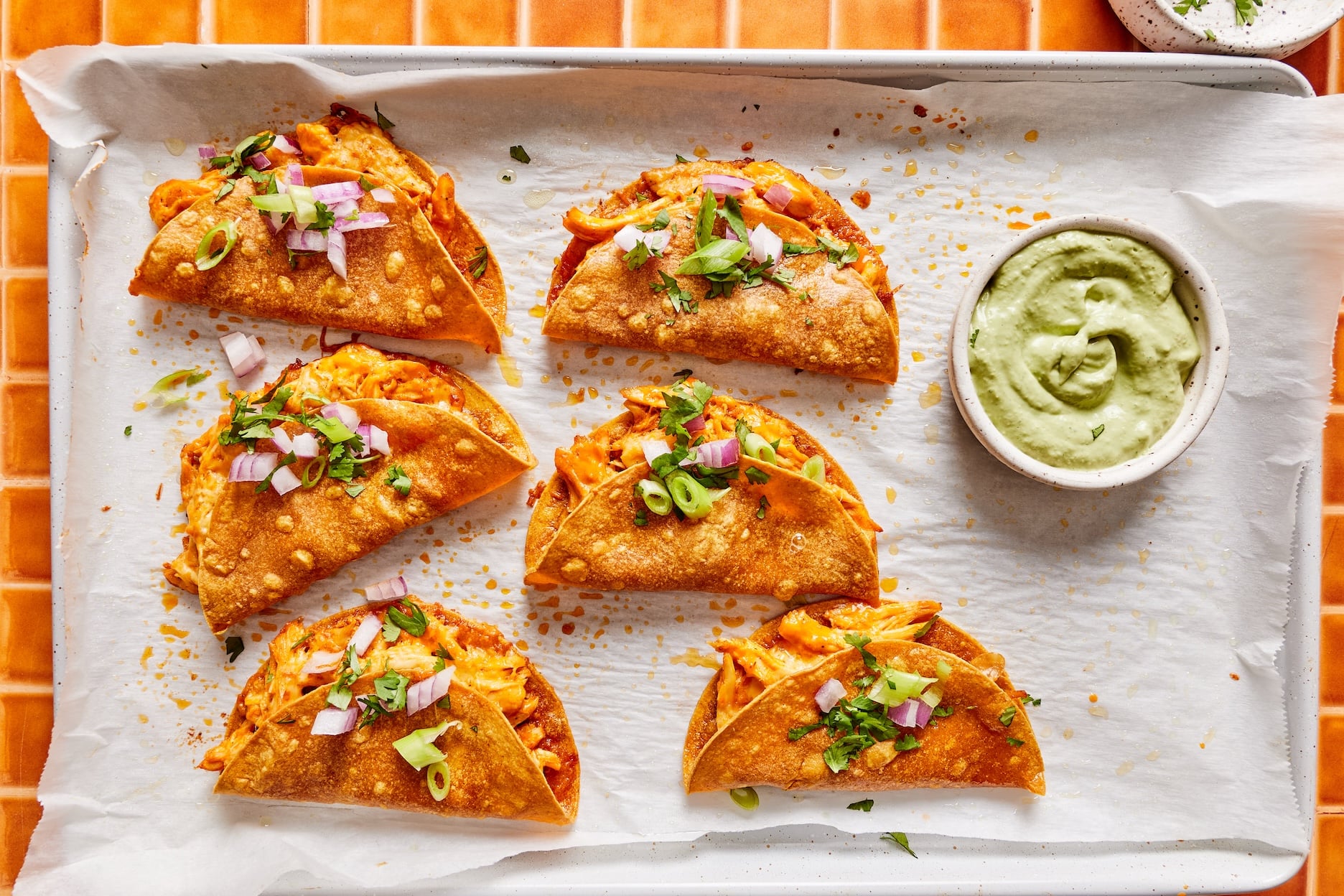 Baked crispy buffalo chicken tacos on a parchment-lined baking sheet garnished with chopped red onion and cilantro with a small bowl of avocado cilantro sauce on the side.